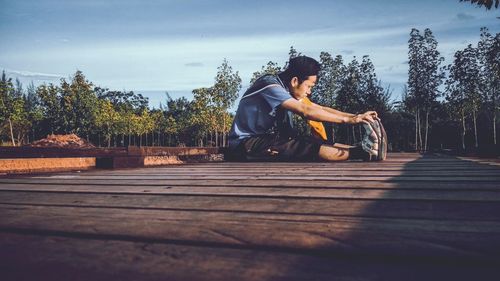 Side view of young man exercising while sitting on boardwalk against sky