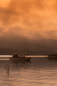 People rowing boat in lake against cloudy sky during sunset