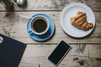 Directly above shot of smart phone with coffee and croissant on wooden table