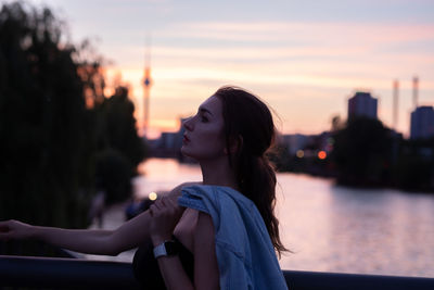 Young woman looking away while standing by railing during sunset