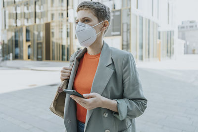 Woman looking away wearing protective face mask using smart phone standing on street in city