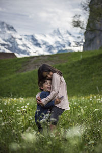 Side view of happy siblings embracing on field against mountain