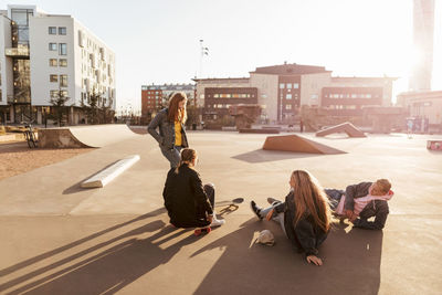 Girl standing while talking to teenage friends sitting at skateboard park during sunny day