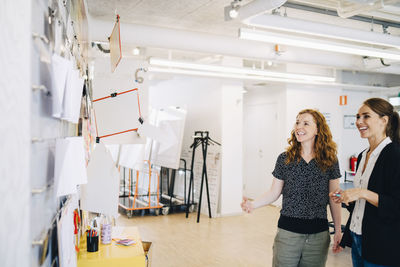 Smiling female colleagues looking at papers on wall at creative office