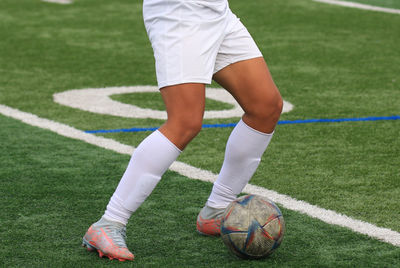 Close up of legs of a young soccer player in a white uniform with the ball and a green turf field.