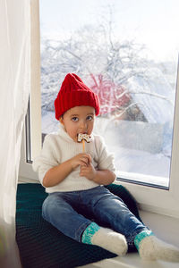 Boy in a white sweater and a red knitted hat sits on the windowsill and eat  gingerbread horse stick