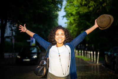 Portrait of smiling young woman with arms raised standing in park