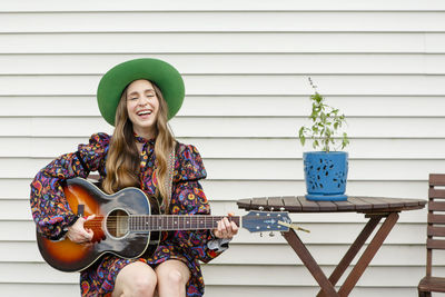 A smiling woman sits at an outdoor table playing acoustic guitar