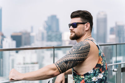 Side view of young man wearing sunglasses standing against railing in city