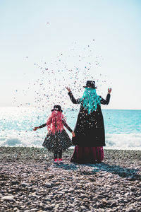 Rear view of mother and daughter standing at beach against clear sky and throwing confettis