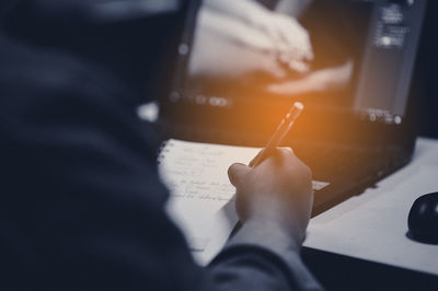 High angle view of man writing on paper while using laptop at desk in office