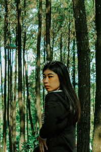 Side view of young woman standing by tree trunk in forest