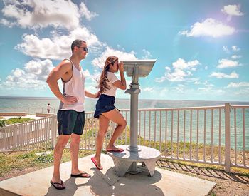 Couple standing by coin-operated binoculars by sea against sky