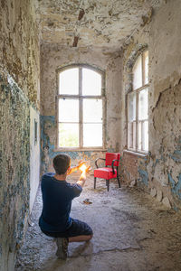 Rear view of young man holding illuminated lighting equipment while kneeling in abandoned house