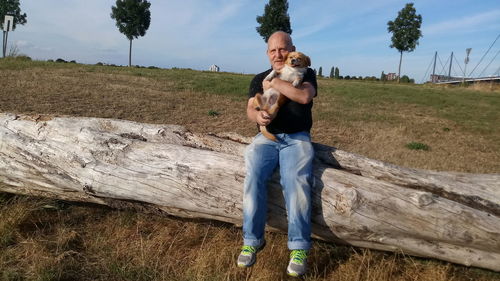 Full length of man carrying dog while sitting on log against sky