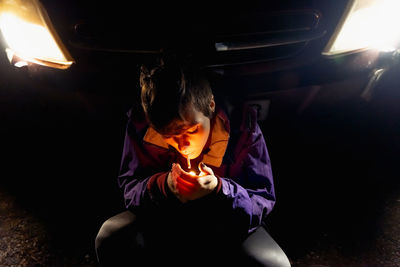 From above provocative confident female in violet and vibrant orange jacket lighting cigarette while sitting alone leaning on automobile with bright headlights at dark night in spain