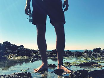 Low section of man standing on rock at beach against clear sky