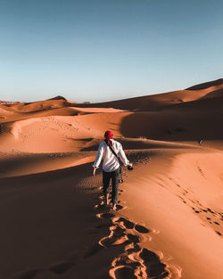 Rear view of man walking on sand dune in desert against clear sky