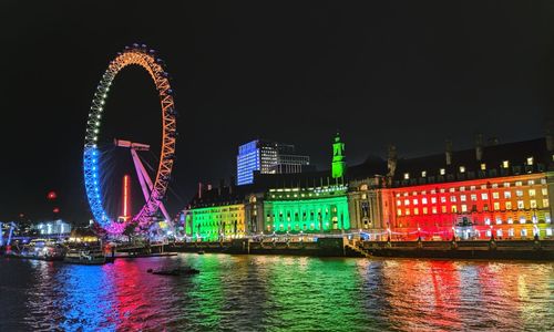 Illuminated ferris wheel by river against buildings at night