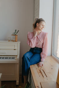 A pretty young woman in jeans and a plaid blouse sits on the windowsill by the window. 