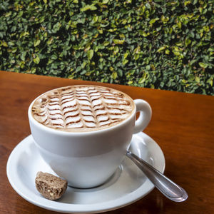 Close-úp coffee cup with decorated shaped latte foam on the wooden table in garden in brazil