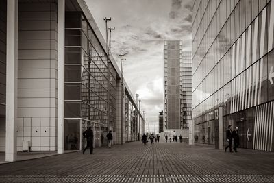 Crowd walking on pathway amidst buildings against cloudy sky