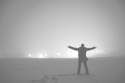 Rear view of man with arms outstretched standing on snow covered field against sky