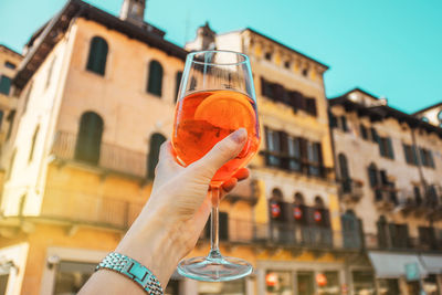 Female hand with glass of orange cocktail spritz near old buildings. sunny day in verona, italy