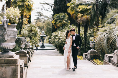 Happy lovers the bride and groom in wedding outfits walk among plants and palm trees in the old park