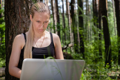 Girl in the forest sits leaning against a tree, working on a laptop. rest at nature. mental health