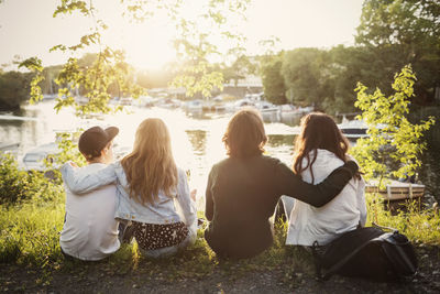 Rear view of teenagers sitting with arms around at lakeshore