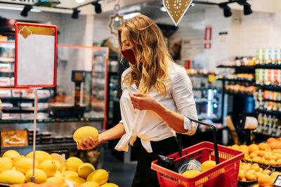 A beautiful young girl in a protective mask with a food basket picks out food on a fruit stand. 