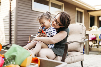 Happy daughter with toys sitting on mother's lap at backyard