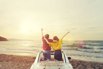 Rear view of couple with arms raised standing in car at beach