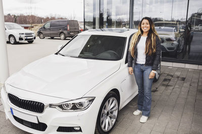 Portrait of smiling young woman standing by car against showroom