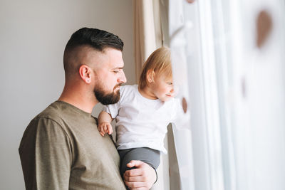 Multinational family, young man father with baby girl on window sill looking at window at home