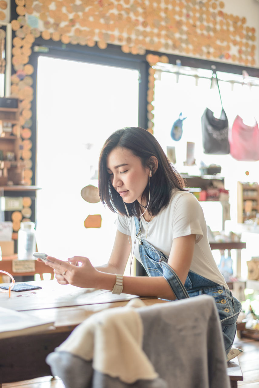YOUNG WOMAN USING PHONE WHILE SITTING ON TABLE IN CAFE