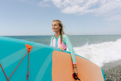 A young athletic blonde woman stands on the beach and holds a paddleboard.