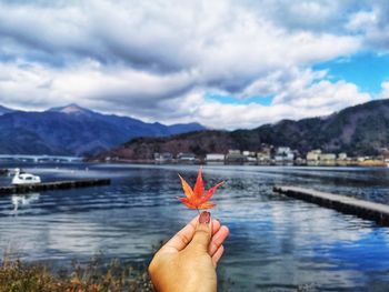 Cropped hand holding autumn leaf by lake against cloudy sky