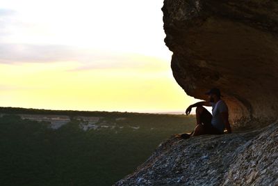 Man sitting on rock formation while looking at landscape during sunset