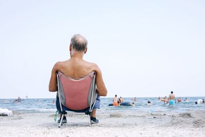 Rear view of shirtless man sitting on camping chair at beach against sky