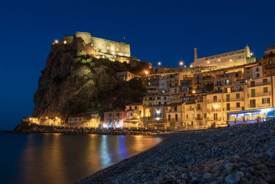 Illuminated buildings by sea against sky at night