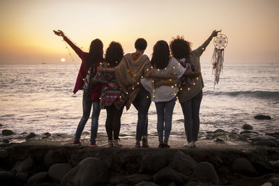 Rear view of friends with illuminated lights standing on shore at beach during sunset