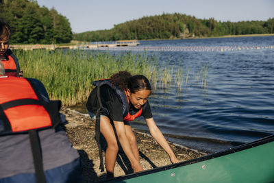 Girl wearing life jacket while holding kayak near lake at summer camp