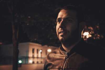 Young man looking away while standing against trees at night