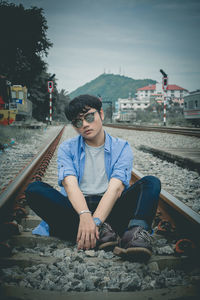 Portrait of young man sitting on railroad track against sky