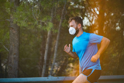 Young man wearing mask jogging against trees