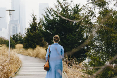 Rear view of young woman with purse wearing trench coat while walking on boardwalk in city