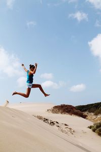 Low angle full length of woman jumping over sand against sky during sunny day