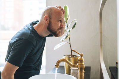 Young adult bearded man looking in mirror in bathroom touching head worried about about hair loss